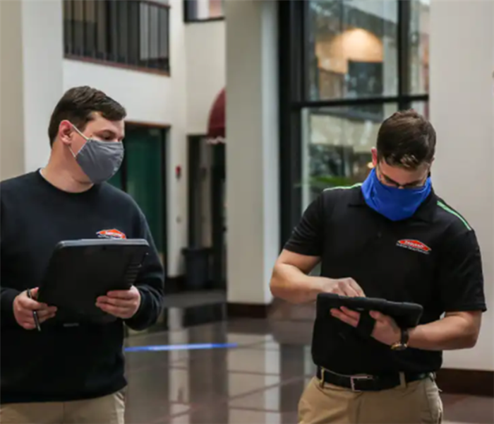 2 men standing inside a building wearing masks and clipboards